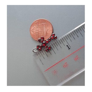 May include: A small, silver cross pendant with red garnet stones. The cross is about 1 inch in length and is shown next to a US penny for size comparison.