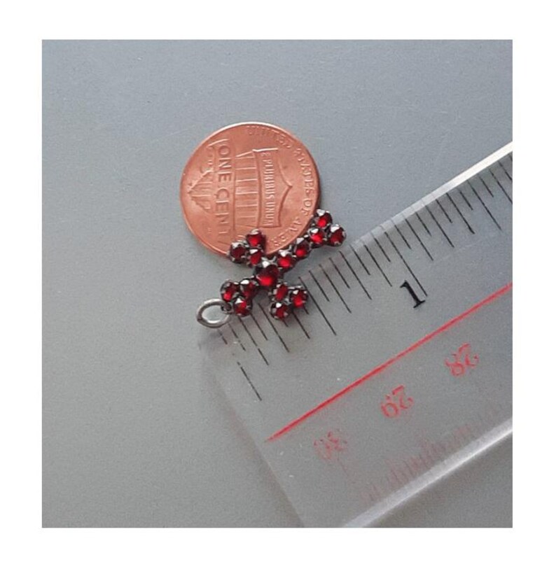 May include: A small, silver cross pendant with red garnet stones. The cross is about 1 inch in length and is shown next to a US penny for size comparison.