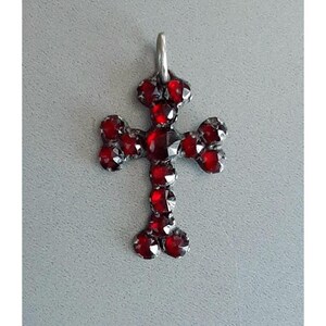 May include: A silver cross pendant with red garnet stones. The cross is set with small, round stones that are arranged in a symmetrical pattern.