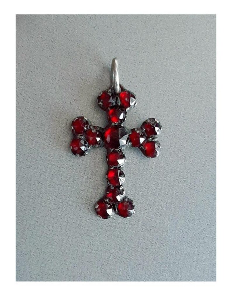 May include: A silver cross pendant with red garnet stones. The cross is set with small, round stones that are arranged in a symmetrical pattern.