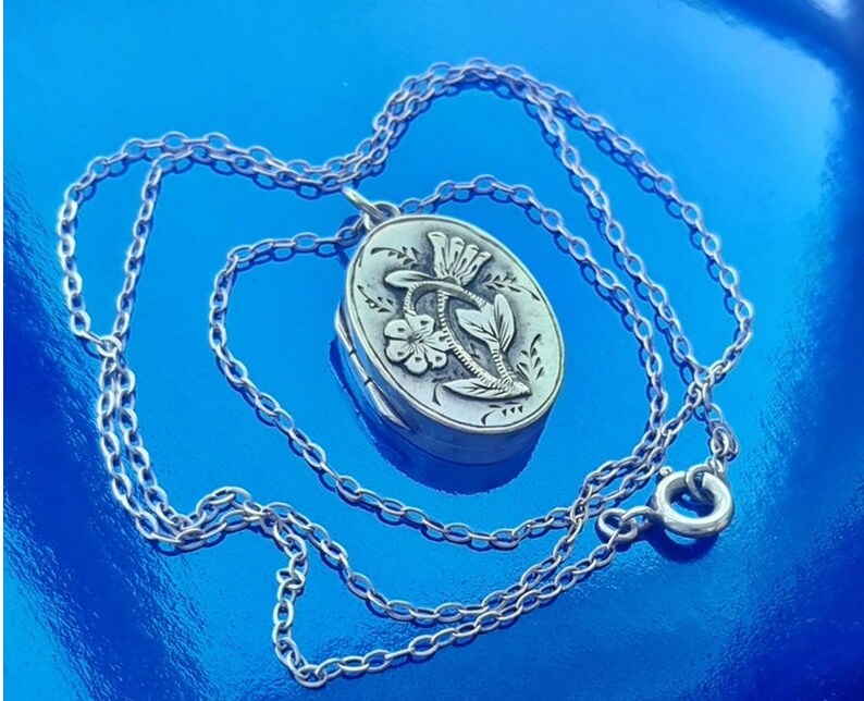 May include: A silver locket necklace featuring a floral design. The locket is oval shaped and has a delicate chain.