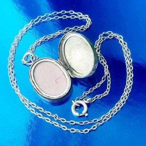 May include: A silver locket necklace with a chain. The locket is oval shaped and has a hinged lid. The locket is open and the inside is visible.