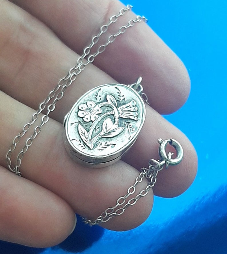 May include: A silver locket necklace featuring a floral design. The locket is oval shaped and intricately engraved with flowers and leaves. The chain is a fine silver chain.