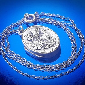 May include: A silver locket necklace featuring a floral design. The locket is oval shaped and has a detailed floral pattern on the front. The chain is silver and has a delicate link design.