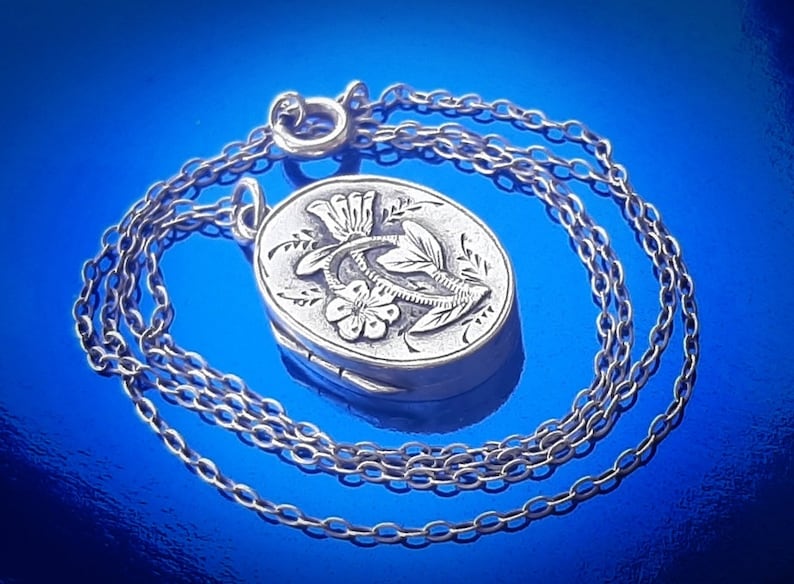May include: A silver locket necklace featuring a floral design. The locket is oval shaped and has a detailed floral pattern on the front. The chain is silver and has a delicate link design.