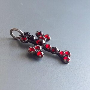 May include: A silver cross pendant with red glass stones. The cross is made of metal and has a loop at the top for hanging.