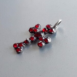 May include: A silver cross pendant with red glass stones. The cross is a simple design with a loop at the top for attaching to a chain.