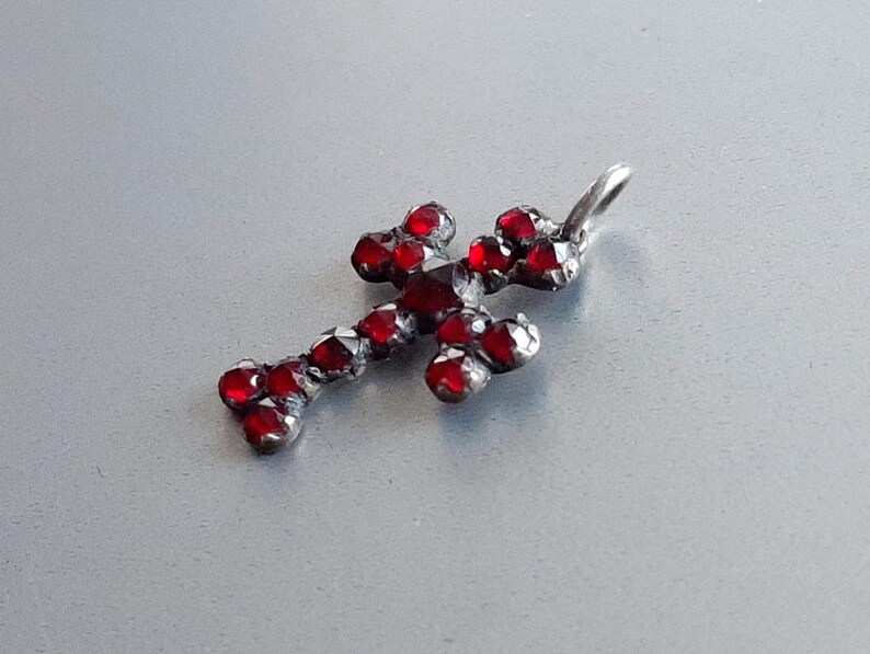 May include: A silver cross pendant with red glass stones. The cross is a simple design with a loop at the top for attaching to a chain.