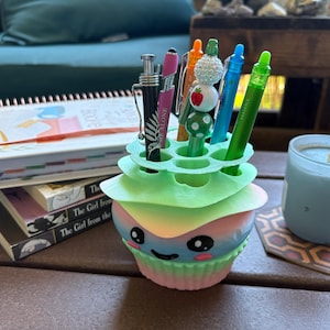 May include: A colorful cupcake-shaped pen holder with a smiling face, holding various pens and pencils. The holder is pastel-colored with a light green top. A candle and books are in the background.