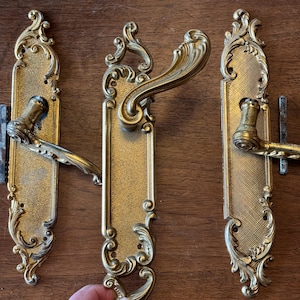May include: Three ornate gold door handles with intricate scrollwork and a decorative design. The handles are on a wooden surface.