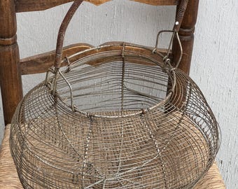 Puede incluir: Una cesta de alambre vintage con forma redondeada y un asa de alambre. La cesta está hecha de alambre de color plateado y tiene un aspecto rústico.