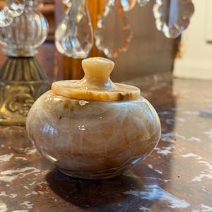 Könnte beinhalten: Ein kleines, rundes Gefäß mit Deckel aus beige-braunem Wirbelstein. Das Gefäß hat eine glatte, polierte Oberfläche und einen kleinen, abgerundeten Knopf auf dem Deckel. Das Gefäß steht auf einer Marmoroberfläche.