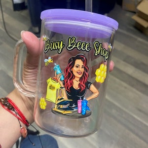 May include: A clear glass tumbler with a purple lid and a straw. The tumbler has a cartoon image of a woman with red hair and a black shirt. The text "Busy Bee Shop" is printed on the tumbler.