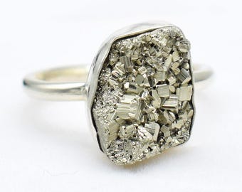 Puede incluir: Un anillo de plata con una gran piedra de pirita de forma irregular. La pirita tiene un aspecto metálico dorado con una superficie texturizada. La banda del anillo es lisa y plateada, contrastando con la piedra rugosa.