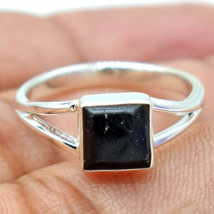 Puede incluir: Un anillo de plata con una piedra cuadrada negra. El anillo tiene un diseño de banda dividida.