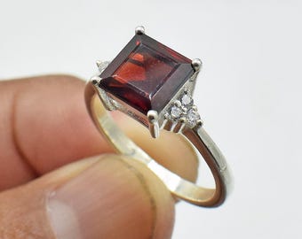 May include: A silver ring with a square red gemstone and smaller clear gemstones on the band.