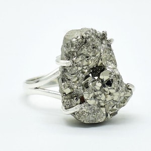Puede incluir: Un anillo de plata con una gran piedra de pirita de forma irregular. La pirita tiene una superficie metálica texturizada con tonos plateados y dorados. La banda del anillo se divide en dos hebras a cada lado.