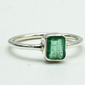 Può includere: Un anello d'argento con una gemma verde rettangolare taglio smeraldo. L'anello ha una fascia sottile e lucida e un'incastonatura a castone che tiene la vibrante pietra verde. L'anello è esposto su una superficie bianca.