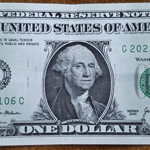 May include: A one dollar bill with a portrait of George Washington. The bill is green and has the text "THE UNITED STATES OF AMERICA" and "ONE DOLLAR" printed on it. The bill also has the serial number "G 20220106 C" printed on it.