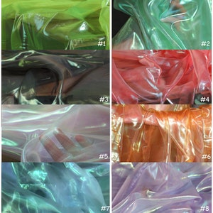Op de afbeelding: Acht stofstalen van iriserende, doorschijnende stof in verschillende kleuren, waaronder groen, zwart, roze, wit, oranje, paars en blauw. De stof is doorschijnend en heeft een glinsterend effect.