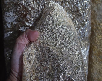 May include: Close-up of a gold metallic fabric with a textured, shimmering surface. The fabric is draped and held in a hand.