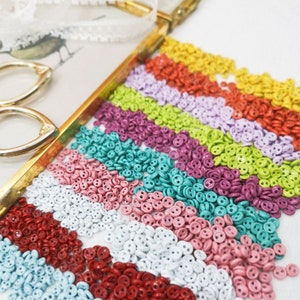 Könnte beinhalten: Sortiment kleiner, zweilöchriger Knöpfe in verschiedenen Farben, darunter Rot, Weiß, Rosa, Blau, Grün, Lila und Gelb. Die Knöpfe sind in Reihen auf einem goldenen Tablett angeordnet.