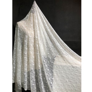May include: Off-white lace fabric with a floral pattern. The delicate material is draped over a form, showcasing its texture and design. The fabric is lightweight and appears to be semi-sheer, with a repeating floral motif.