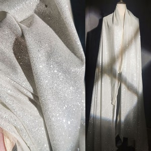 Puede incluir: Primer plano y plano completo de una tela blanca brillante. La tela tiene una textura delicada, similar a una red, y está cubierta de purpurina brillante. La tela está drapeada sobre un maniquí, mostrando su fluidez y caída.