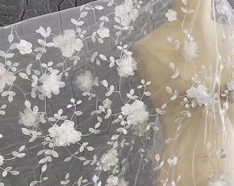 May include: White floral lace fabric with delicate 3D flowers and leaves. The fabric is sheer and lightweight, perfect for creating garments, accessories, or home decor.
