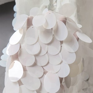 May include: White fabric with a pattern of overlapping, round sequins. The sequins are arranged in a fish scale pattern.