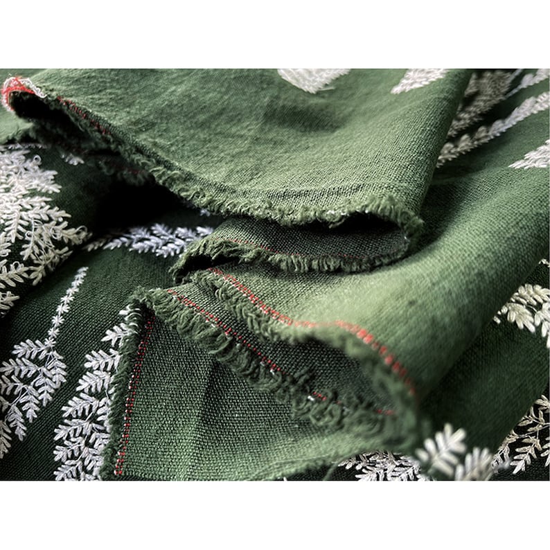 May include: Close-up of a dark green fabric with a white embroidered fern pattern. The fabric has a raw edge and is folded over itself.