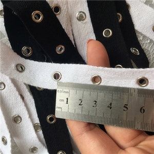 K&ouml;nnte beinhalten: Schwarzes und wei&szlig;es Baumwollband mit silbernen &Ouml;sen. Das Band wird mit einem Lineal gemessen, das 6 Zentimeter anzeigt.