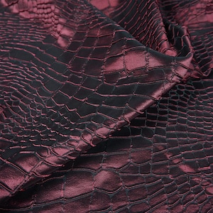 May include: A close-up shot of a fabric with a crocodile skin pattern. The material is a deep burgundy with black shading, giving a shimmering appearance. The fabric is folded, highlighting the texture and sheen of the material.