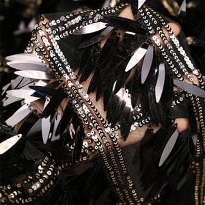 Puede incluir: Primer plano de una tela negra con lentejuelas doradas, plateadas y negras en forma de hoja. Las lentejuelas están cosidas sobre una tela de malla negra transparente.