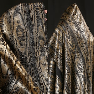 May include: A close-up of a fabric with a gold and black pattern. The fabric has a vertical striped design with a metallic sheen. The material is draped, showing the texture and flow of the fabric.