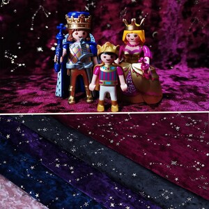 Puede incluir: Una familia real de tres figuritas, un rey, una reina y un príncipe, de pie sobre una tela de terciopelo burdeos con un patrón de estrellas doradas. El rey lleva una capa azul y una armadura dorada, la reina lleva un vestido dorado con un corpiño rosa y el príncipe lleva una camisa a rayas rojas y blancas y pantalones dorados.