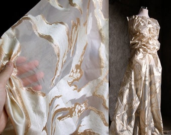 May include: White sheer fabric with a gold metallic swirl pattern. The fabric is draped over a hand and also shown as a dress.