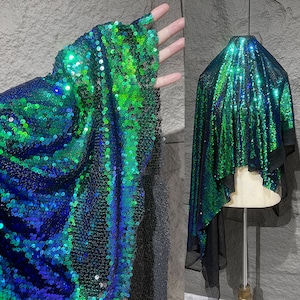 Op de afbeelding: Een glinsterende stof met pailletten in groen- en blauwtinten. De stof is over een vorm gedrapeerd, waardoor de textuur en kleurvariaties zichtbaar worden. De pailletten vangen het licht op en creëren een schitterend effect, ideaal voor avondkleding.