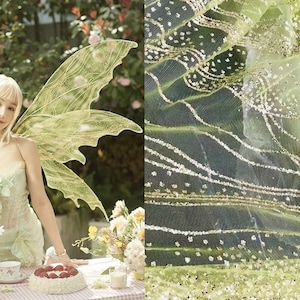 May include: A woman wearing a green fairy costume with large, green wings, holding a plate of strawberries and whipped cream. The background is a garden with flowers and greenery.