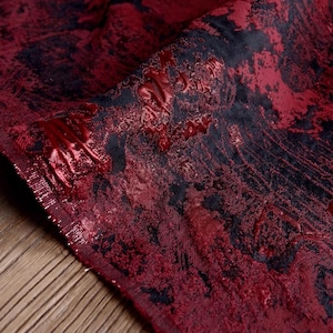 May include: Close-up of a textured fabric with a deep red and black pattern. The fabric has a shiny, almost wet appearance in areas, with a raised, embossed design. The overall effect is rich and luxurious, suitable for apparel or home decor.