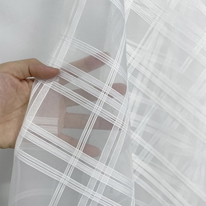 以下が含まれることがあります： 格子模様の白いシアー生地。生地は持ち上げられ、その透明性が明らかになっています。格子は白い線で形成され、テクスチャ効果を生み出しています。カーテンや装飾目的などに適した、軽くて繊細な素材に見えます。