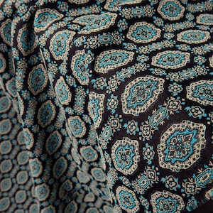 Puede incluir: Primer plano de una tela oscura con un patrón repetitivo de formas tipo paisley turquesa y blanquecino sobre un fondo negro. La tela está doblada, mostrando la textura y los detalles del diseño. El efecto general es un textil rico y detallado.