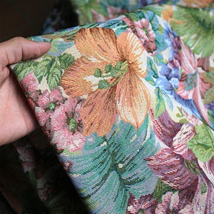 May include: Close-up of a colorful floral fabric with a tapestry-like texture. The fabric features a variety of flowers, including pink, orange, and blue, as well as green leaves.