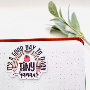 May include: A white sticker with a rainbow, pencil, and apple design. The text reads "It's a good day to teach tiny humans".