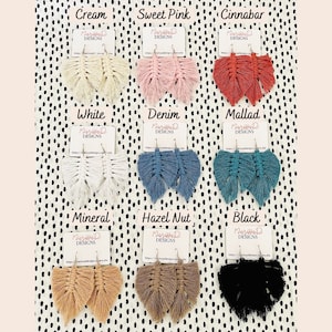 May include: Nine pairs of handmade macrame earrings in various colors, including cream, sweet pink, cinnabar, white, denim, mallad, mineral, hazel nut, and black. Each pair is displayed on a white card with the brand name "Maritta D Designs" and a website address.