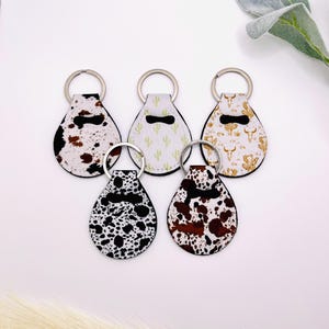 May include: Five keychains with different patterns. The keychains are shaped like teardrops and have silver rings. The patterns include cowhide, cactus, and a southwestern design.
