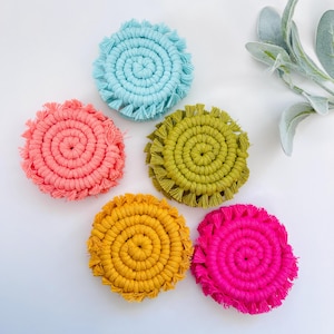 May include: Five colorful round crocheted coasters with fringe. The coasters are in shades of blue, pink, green, yellow, and pink.