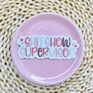 Pu&ograve; includere: Un adesivo bianco con il testo "Shit Show Supervisor" in lettere rosa, rosse e marroni con accenti di glitter.