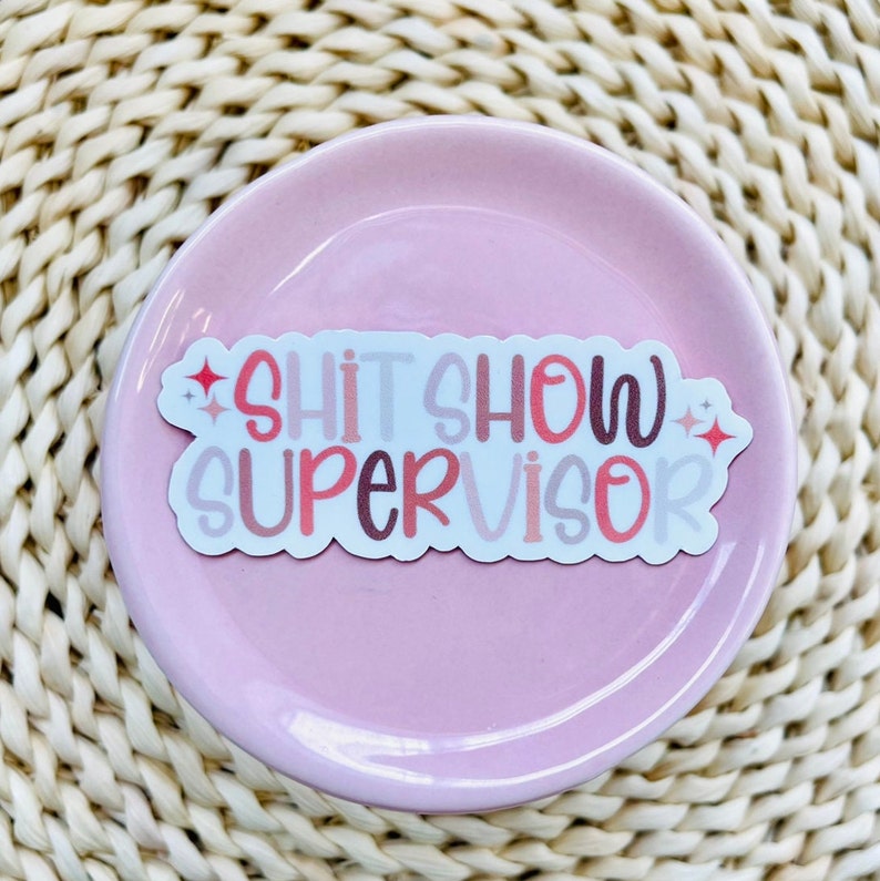 Pu&ograve; includere: Un adesivo bianco con il testo "Shit Show Supervisor" in lettere rosa, rosse e marroni con accenti di glitter.