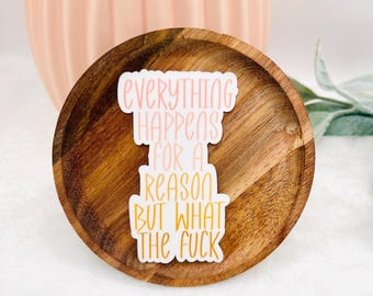 May include: A white sticker with the text "Everything Happens For A Reason But What The Fuck" in pink and yellow lettering. The sticker is on a wooden plate with a pink vase in the background.